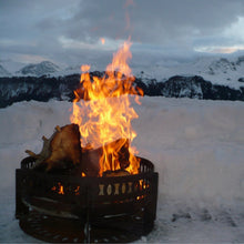 Laden Sie das Bild in den Galerie-Viewer, Ethno Ø 75 cm Design Feuerschale im Winter mit großen Holzstücken und Feuer. Feuerschale steht auf einem Feuerschalendeckel mit dem Durchmesser 75 cm
